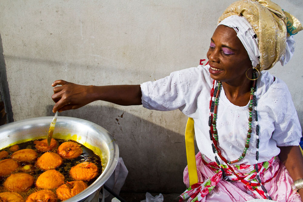 Woman frying orange food in a large pot, wearing a headwrap and colorful clothes.