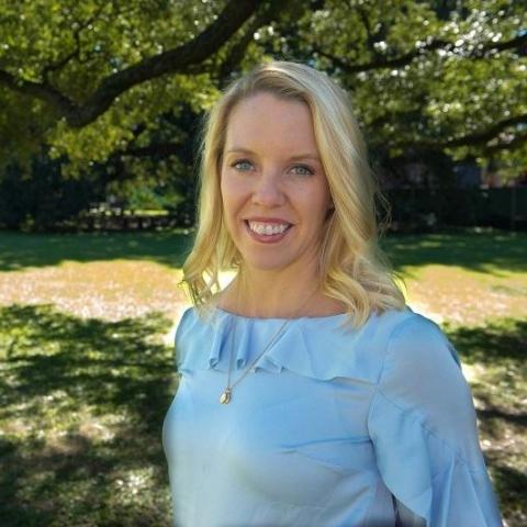 A blonde woman in a blue blouse poses in front of greenery.