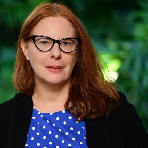 A red-haired woman in a blue polka dot shirt and glasses poses in front of greenery.