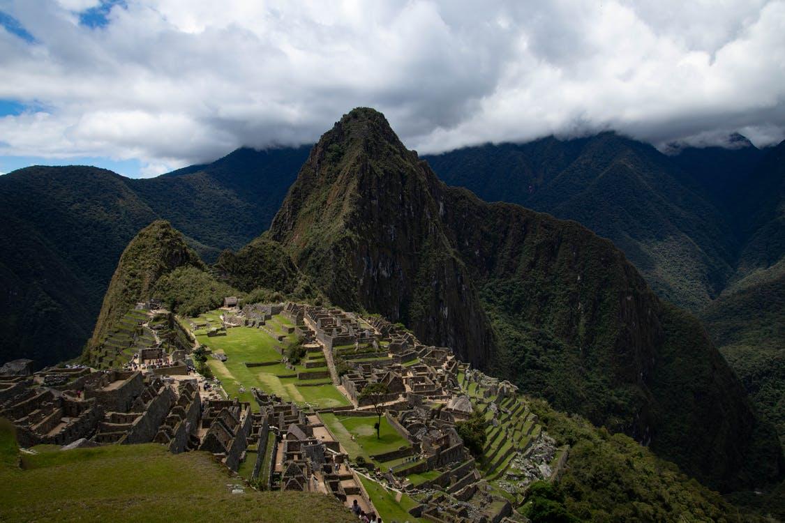 Image of green, mountainous landscape covered with terraced stone ruins. There is a peaked rocky mountaintop in the distance, white clouds, and a grey sky.