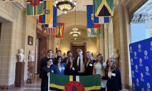 Group of formally dressed young adults holding the Dominica flag in a hall with international flags.