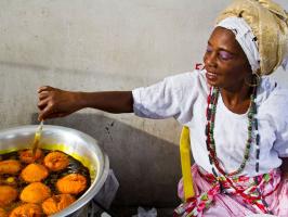Woman frying orange food in a large pot, wearing a headwrap and colorful clothes.