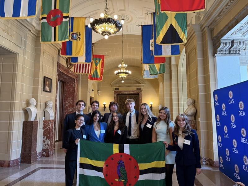 Group of formally dressed young adults holding the Dominica flag in a hall with international flags.